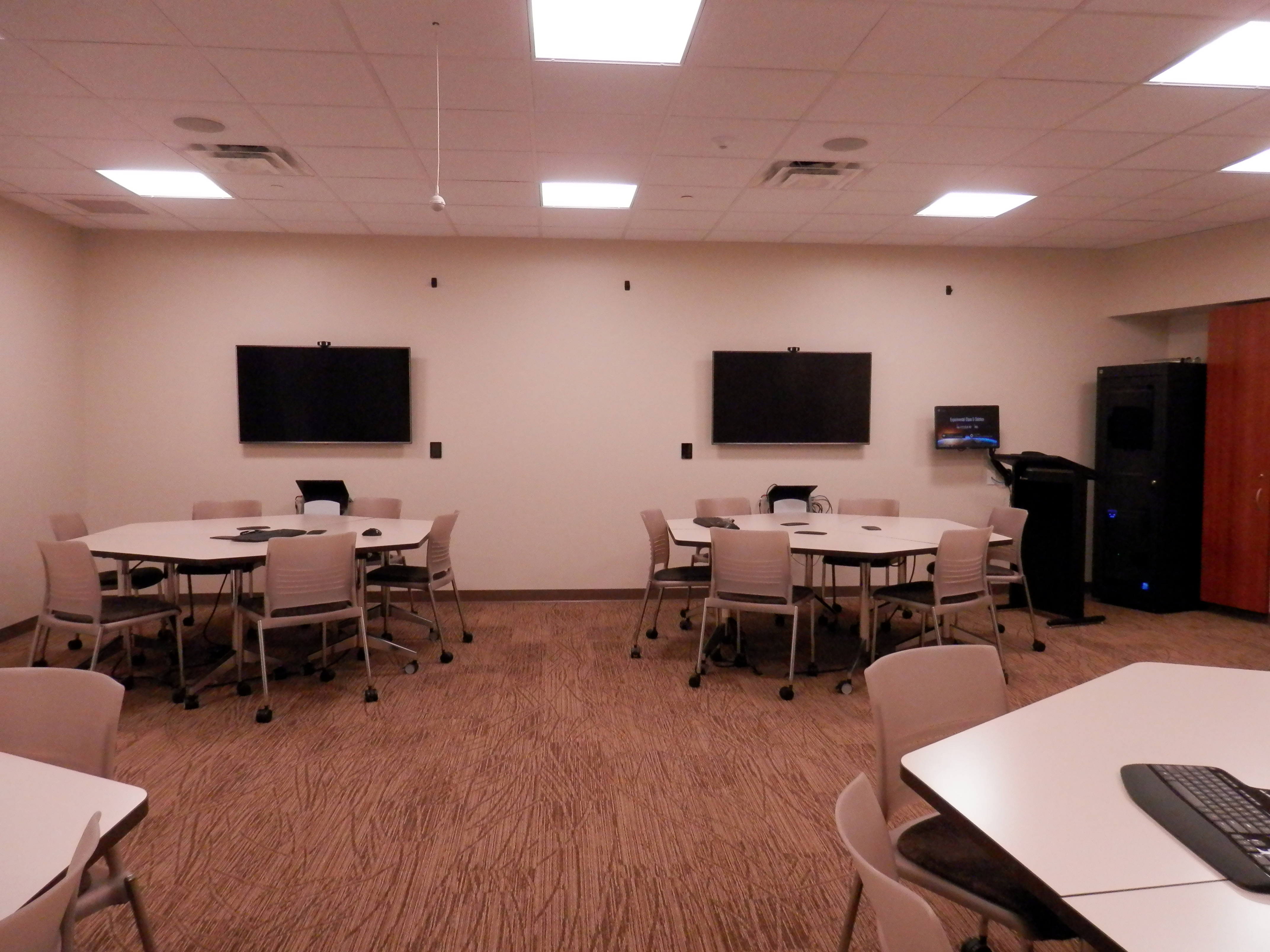 Photo partially reveals the inside of the experimental classroom. There are one ceiling speaker, four 6-seat configurable tables with a monitor on the wall behind each table, one instructor stand and the control closet.