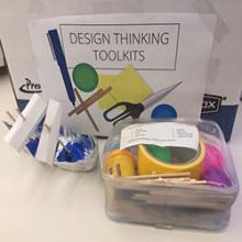 On a table is a clear, plastic case filled with craft supplies, visibly including duct tape and ping pong balls. Beside the case is a boat made of aluminum foil and paper. In the background is a large cardboard box labeled “Design Thinking Toolkits.”