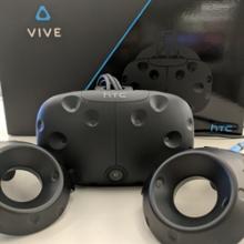 A headset and two controllers of a HTC Vive VR system are placed in front of the packaging box.